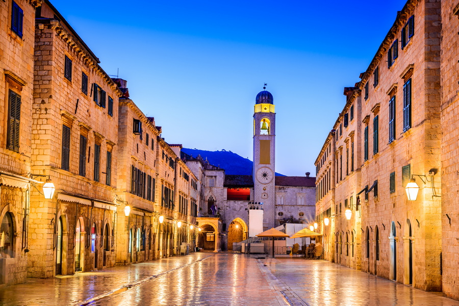 A cobblestone street in Dubrovnik, Croatia, with tall buildings on either side and a church tower in the distance.