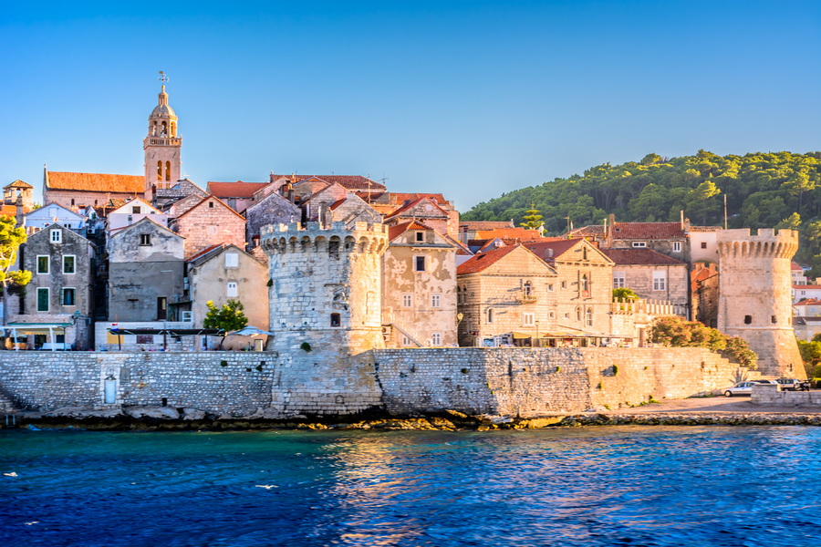 A panoramic view of Korčula's Old Town, showcasing its medieval architecture, the city walls, and the vibrant waterfront scene with people strolling along the promenade.