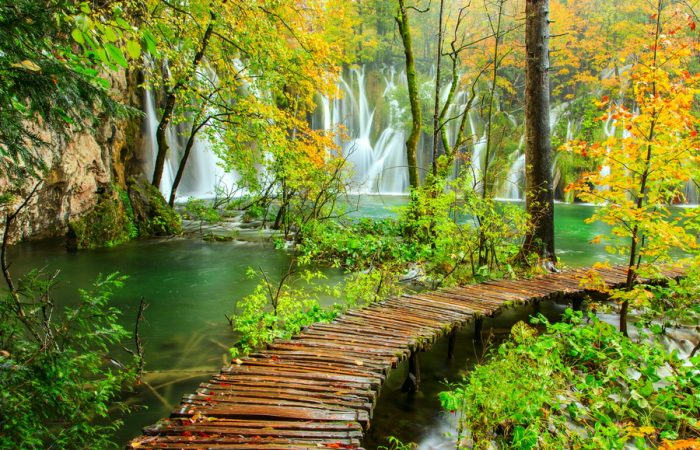 A wooden path winding through a lush autumn forest towards a cascading waterfall in Plitvice Lakes National Park, Croatia.