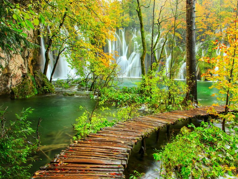 A wooden path winding through a lush autumn forest towards a cascading waterfall in Plitvice Lakes National Park, Croatia.