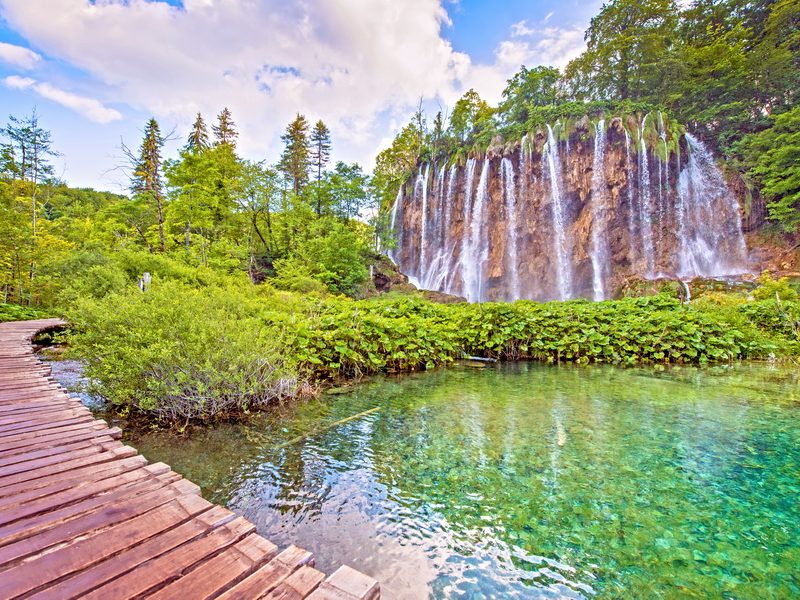 A wooden boardwalk winding through a lush forest landscape, leading to a stunning waterfall cascading into a turquoise lake in Plitvice Lakes National Park, Croatia.