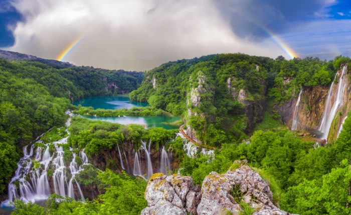 A panoramic view of Plitvice Lakes National Park, featuring multiple waterfalls, lush forests, and a double rainbow.