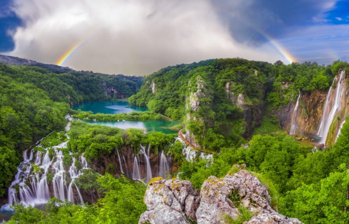 A panoramic view of Plitvice Lakes National Park, featuring multiple waterfalls, lush forests, and a double rainbow.