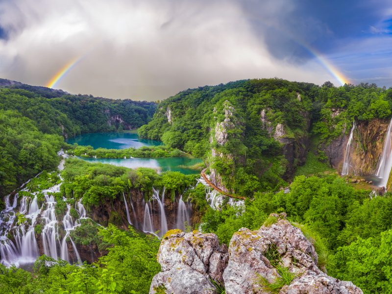 A panoramic view of Plitvice Lakes National Park, featuring multiple waterfalls, lush forests, and a double rainbow.