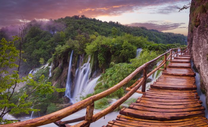 A wooden walkway winding through a lush forest with stunning waterfalls in the background at Plitvice Lakes National Park, Croatia.