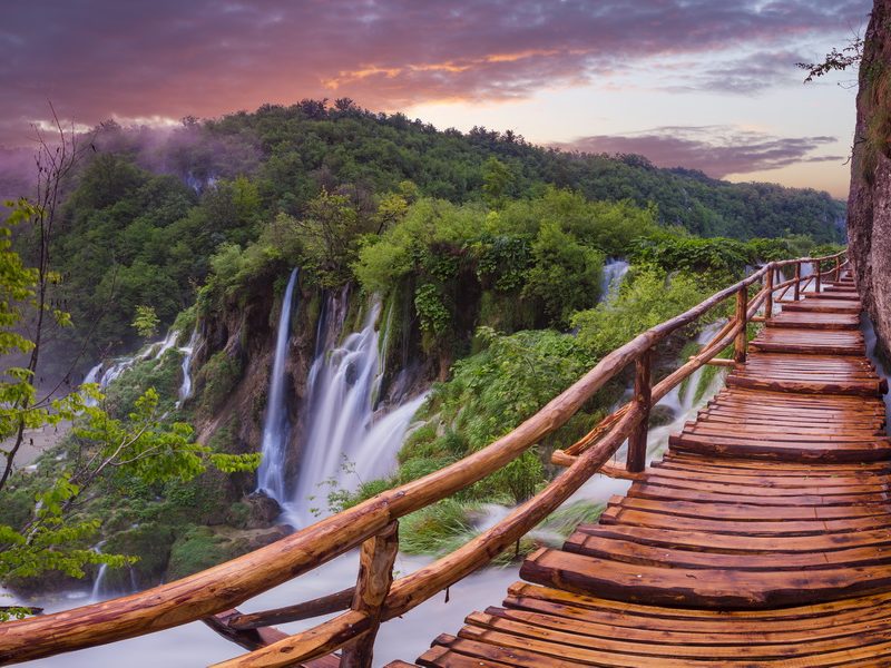 A wooden walkway winding through a lush forest with stunning waterfalls in the background at Plitvice Lakes National Park, Croatia.