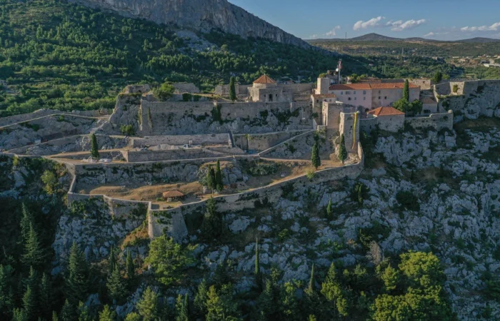 Dramatic aerial photo of Klis Fortress perched atop rocky cliffs with a walled complex and red-roofed buildings, surrounded by lush hills in Dalmatia, Croatia.
