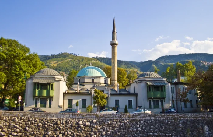 View of Sarajevo’s Careva (Emperor’s) Mosque with turquoise dome and slender minaret, set against green hills along the Miljacka River in Bosnia and Herzegovina.
