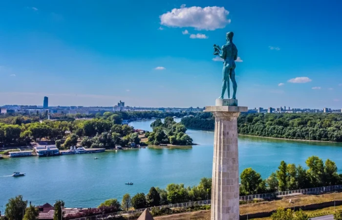 Panoramic view of Belgrade’s Victor Monument at Kalemegdan Fortress, overlooking the confluence of the Sava and Danube rivers under a blue sky.