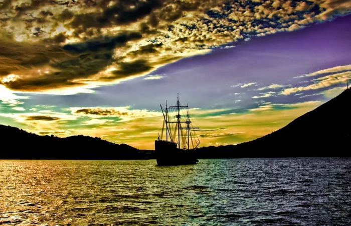 Silhouette of a tall sailing ship at sunset, dramatic clouds and colorful sky reflected on calm waters, evoking exploration and maritime adventure.