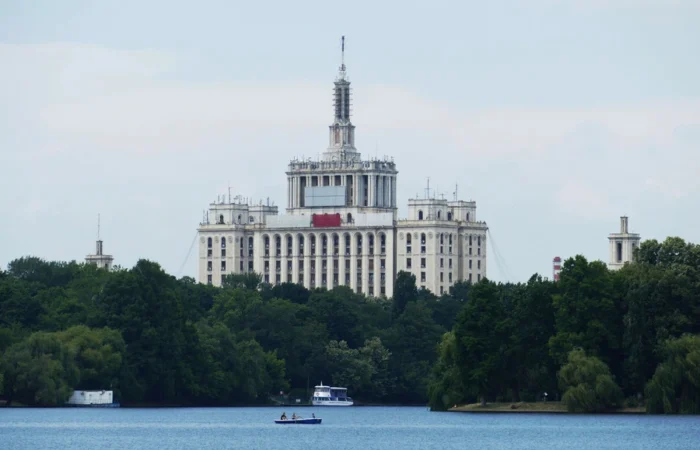 View across Herăstrău Lake towards the imposing Soviet-style House of the Free Press in Bucharest, Romania, framed by green trees and a calm waterfront.