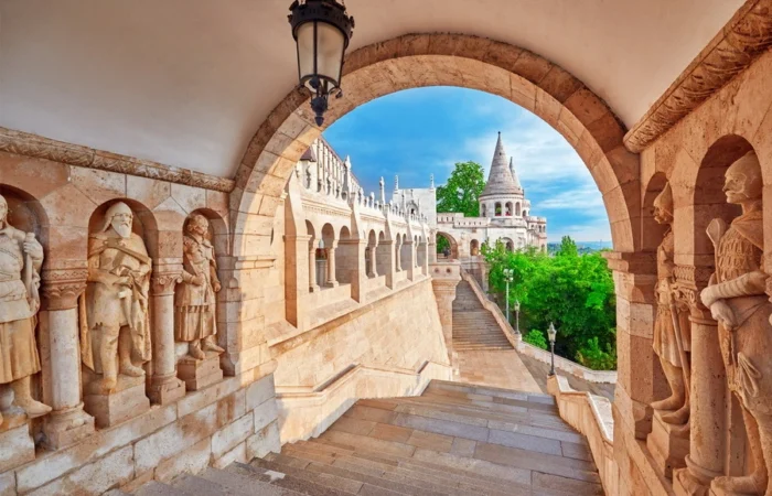 Stone archway and historic statues of Hungarian figures leading to the turrets and terraces of Fisherman’s Bastion, with panoramic views over Budapest and the Danube.