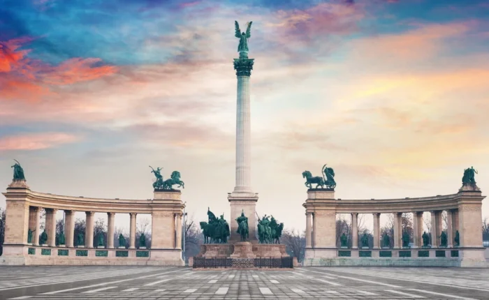 Heroes’ Square in Budapest, Hungary, featuring the iconic Millennium Monument with statues of national leaders, horses, and the angel-topped column beneath a colorful evening sky.