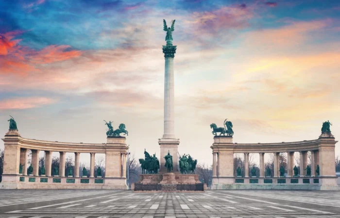 Heroes’ Square in Budapest, Hungary, featuring the iconic Millennium Monument with statues of national leaders, horses, and the angel-topped column beneath a colorful evening sky.