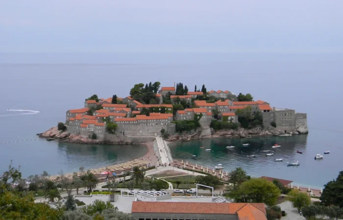 Elevated view of Sveti Stefan Island’s stone villas with orange roofs, the connecting sandy causeway, calm Adriatic waters, and lush surrounding landscape near Budva, Montenegro.