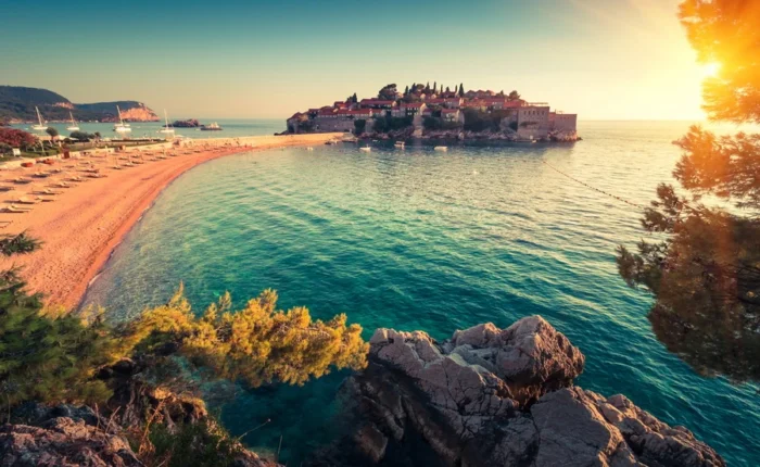 Idyllic view of Sveti Stefan islet and its historic hotel complex, pink sandy beach, sun loungers, turquoise Adriatic Sea, and pine trees at sunset near Budva, Montenegro.