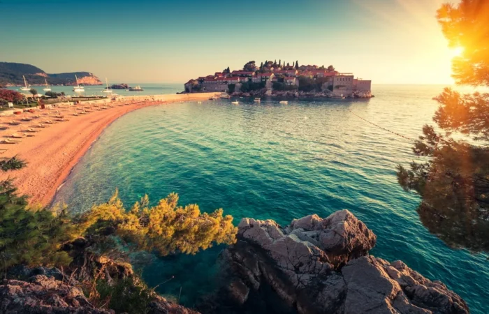 Idyllic view of Sveti Stefan islet and its historic hotel complex, pink sandy beach, sun loungers, turquoise Adriatic Sea, and pine trees at sunset near Budva, Montenegro.