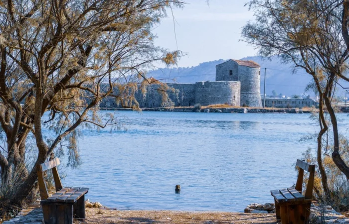 Stone fortress ruins of Butrint framed by trees, seen across tranquil blue water with benches in the foreground, in southern Albania.