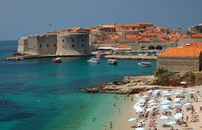 Tourists enjoying the sun at Banje Beach with umbrellas and swimmers, boats on emerald water, and the medieval stone walls and orange rooftops of Dubrovnik Old Town in the background.