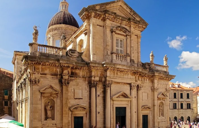 Majestic Baroque Dubrovnik Cathedral with statues, ornate facade, and a grand domed roof against a clear blue sky, surrounded by Old Town’s historic streets.