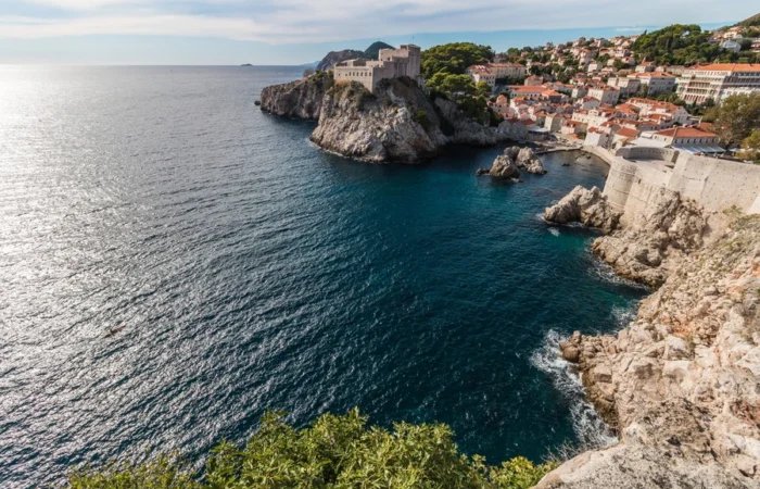 Aerial view of Lovrijenac Fortress perched on rocky cliffs above deep blue Adriatic waters, with Dubrovnik’s old town and red rooftops stretching along the coastline.