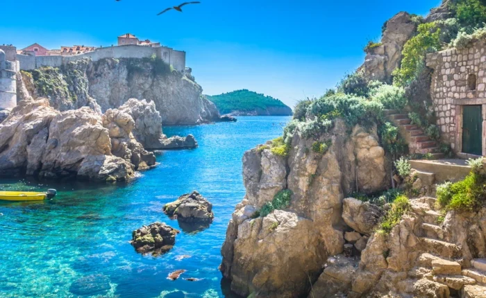 Sunlit Adriatic cove beneath Dubrovnik’s medieval city walls, with rocky cliffs, stone steps, lush plants, crystal-clear turquoise water, and birds soaring on a bright day.
