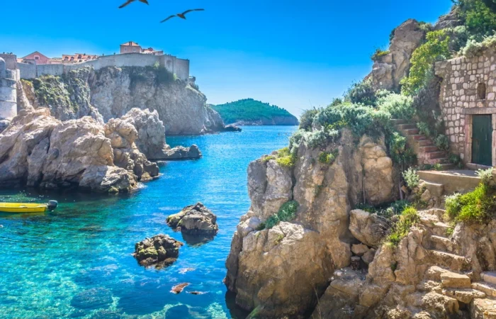Sunlit Adriatic cove beneath Dubrovnik’s medieval city walls, with rocky cliffs, stone steps, lush plants, crystal-clear turquoise water, and birds soaring on a bright day.
