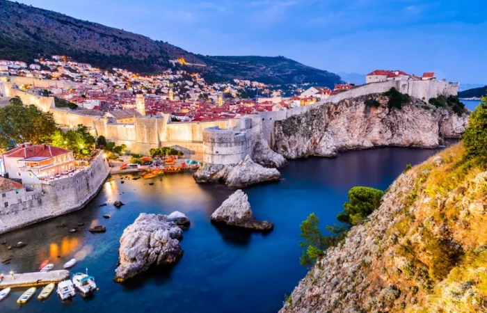 Illuminated Dubrovnik city walls and bastions glowing at sunset, with small boats in the harbor, huge stone cliffs, and hillside houses overlooking the Adriatic Sea.