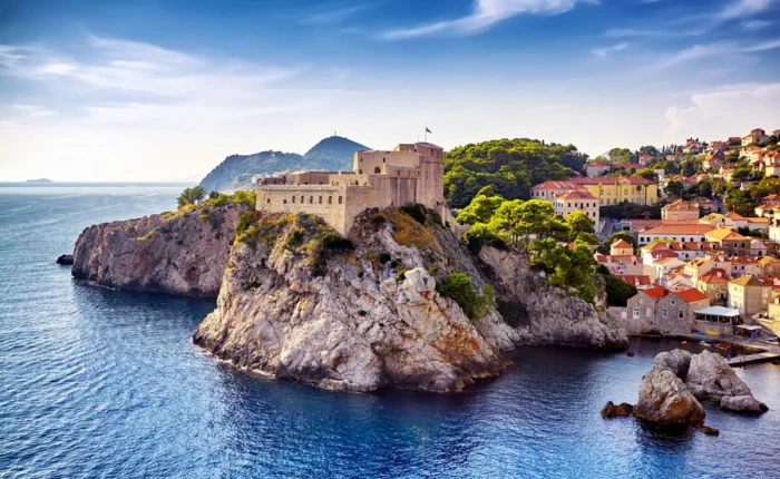 Lovrijenac Fortress perched on rocky cliffs above the Adriatic Sea, surrounded by pine trees and old town buildings, with a bright sky over Dubrovnik, Croatia.