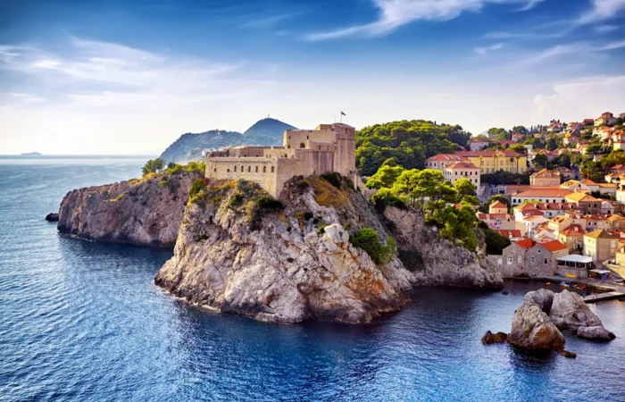 Lovrijenac Fortress perched on rocky cliffs above the Adriatic Sea, surrounded by pine trees and old town buildings, with a bright sky over Dubrovnik, Croatia.