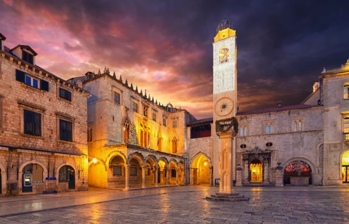 Evening view of Luža Square in Dubrovnik’s Old Town, with the iconic medieval clock tower illuminated and Renaissance palaces glowing under dramatic twilight clouds.