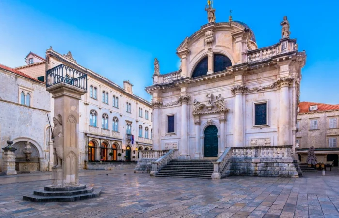 Dubrovnik’s Baroque Church of St. Blaise with grand steps, statues, and adjacent medieval Orlando’s Column in a lively Old Town square beneath a clear blue sky.