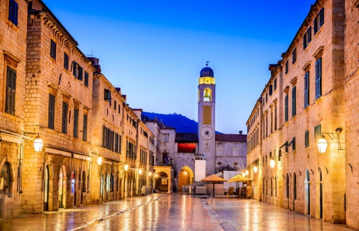 Glistening Stradun main street lined with golden stone buildings, warm streetlights, and the illuminated Clock Tower at twilight in Dubrovnik Old Town, Croatia.