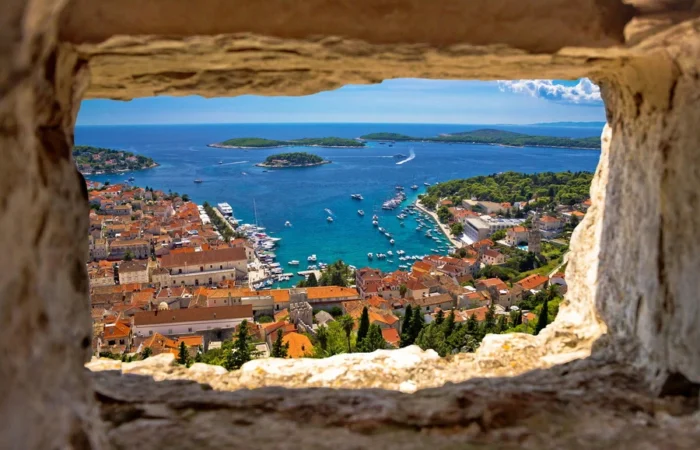 Spectacular view of Hvar Town’s terracotta rooftops, marina full of boats, and the blue Adriatic dotted with Pakleni Islands, seen through an old fortress window.