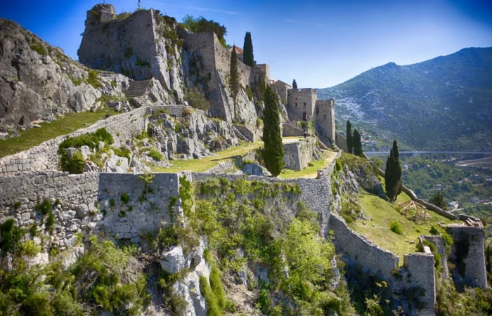 Panoramic view of the ancient stone walls and dramatic ramparts of Klis Fortress clinging to a rocky ridge, with cypress trees and Dalmatian mountains in the background.