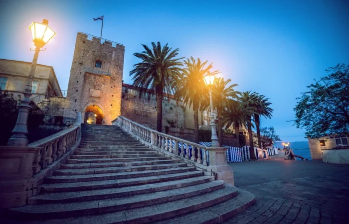 Stone staircase leading to Revelin Tower, Land Gate entrance of Korčula Old Town, Croatia, illuminated at dusk with palm trees and historic architecture.
