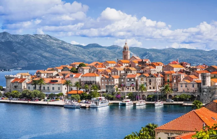 Panoramic view of Korčula Old Town, Croatia, with waterfront marina, historic stone walls, red-roofed houses, and St. Mark’s Cathedral belltower against the backdrop of Pelješac mountains.
