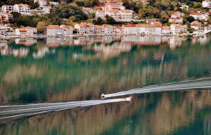 Waterfront houses with scenic reflections in Kotor Bay, Montenegro, tranquil Adriatic water, boats making trails, and lush green hillside.