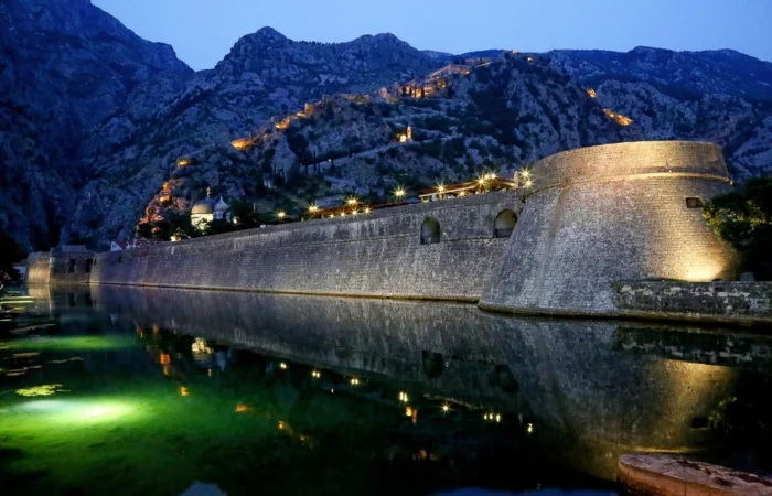 Illuminated medieval city walls and fortress of Kotor, Montenegro, at night, reflected in water, with dramatic mountains in the background.