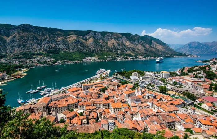 Panoramic aerial view of Kotor Old Town, Montenegro, with red-tiled roofs, marina, cruise ships, blue bay, and dramatic mountains in the background.