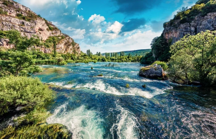 Stunning river canyon with turquoise water, cascades, lush greenery, and rocky cliffs in Krka National Park, Croatia, under a vibrant sky.
