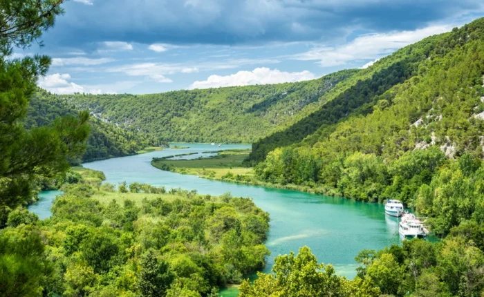 Scenic panorama of the Krka River winding through lush green forests in Krka National Park, Croatia, with excursion boats and dramatic hills under a blue sky.