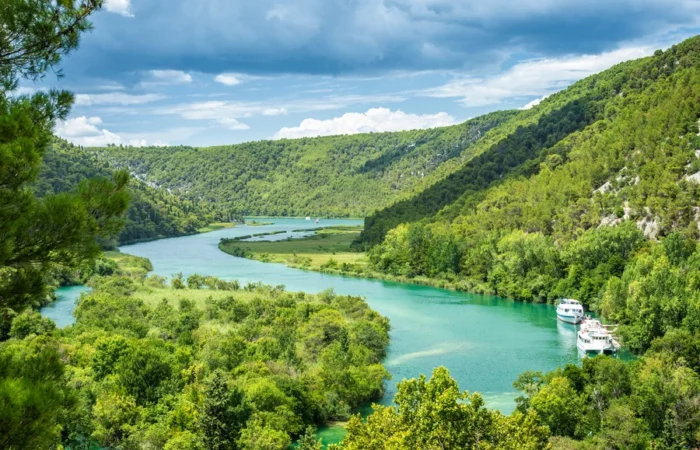 Scenic panorama of the Krka River winding through lush green forests in Krka National Park, Croatia, with excursion boats and dramatic hills under a blue sky.