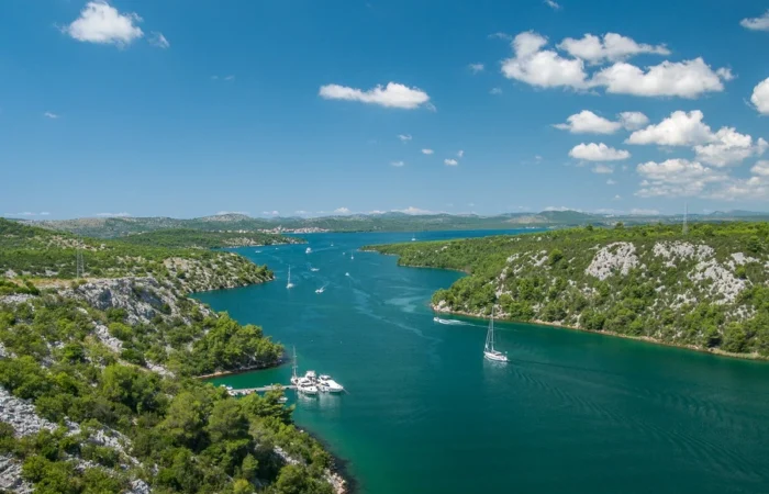 Panoramic view of yachts and sailboats cruising the turquoise Krka River, Croatia, surrounded by lush green hills and rocky landscapes under a bright summer sky.