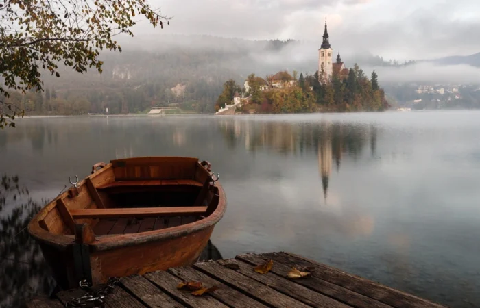 Traditional wooden pletna boat on the calm waters of Lake Bled with the iconic Bled Island and Church of the Assumption shrouded in morning mist, surrounded by forested hills, Slovenia.