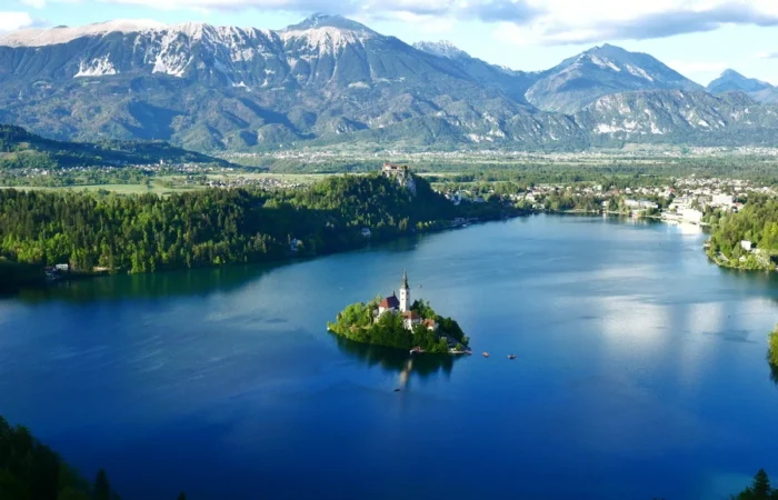 Clear panoramic view of Lake Bled with its central island church, emerald forests, sparkling blue water, and the dramatic snow-capped Julian Alps in the background, Slovenia.