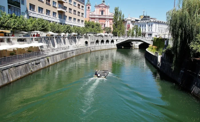 Daytime view of Ljubljanica River and Triple Bridge in Ljubljana, Slovenia, with boat, riverside cafes, and the pink Franciscan Church of the Annunciation.
