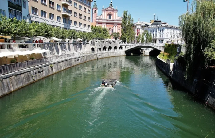 Daytime view of Ljubljanica River and Triple Bridge in Ljubljana, Slovenia, with boat, riverside cafes, and the pink Franciscan Church of the Annunciation.
