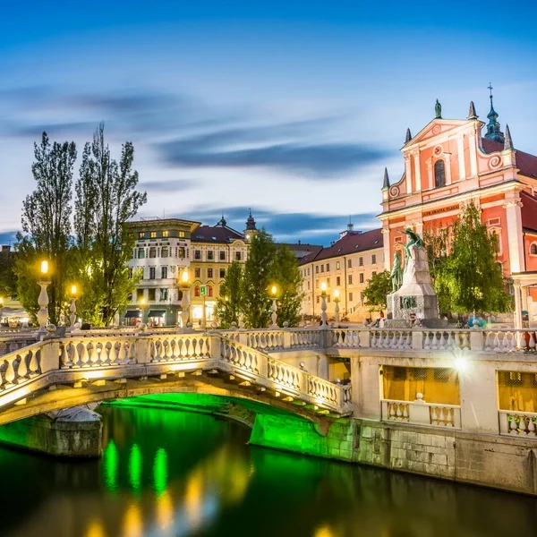 Illuminated Triple Bridge and Prešeren Square at blue hour in Ljubljana, Slovenia, with the pink Franciscan Church of the Annunciation and historic city center.