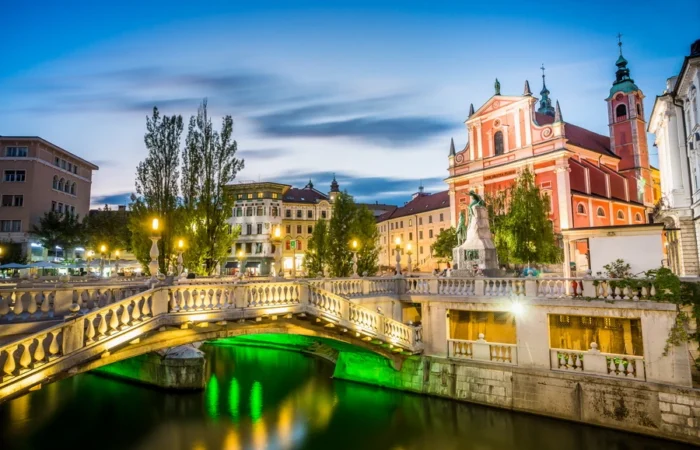 Illuminated Triple Bridge and Prešeren Square at blue hour in Ljubljana, Slovenia, with the pink Franciscan Church of the Annunciation and historic city center.